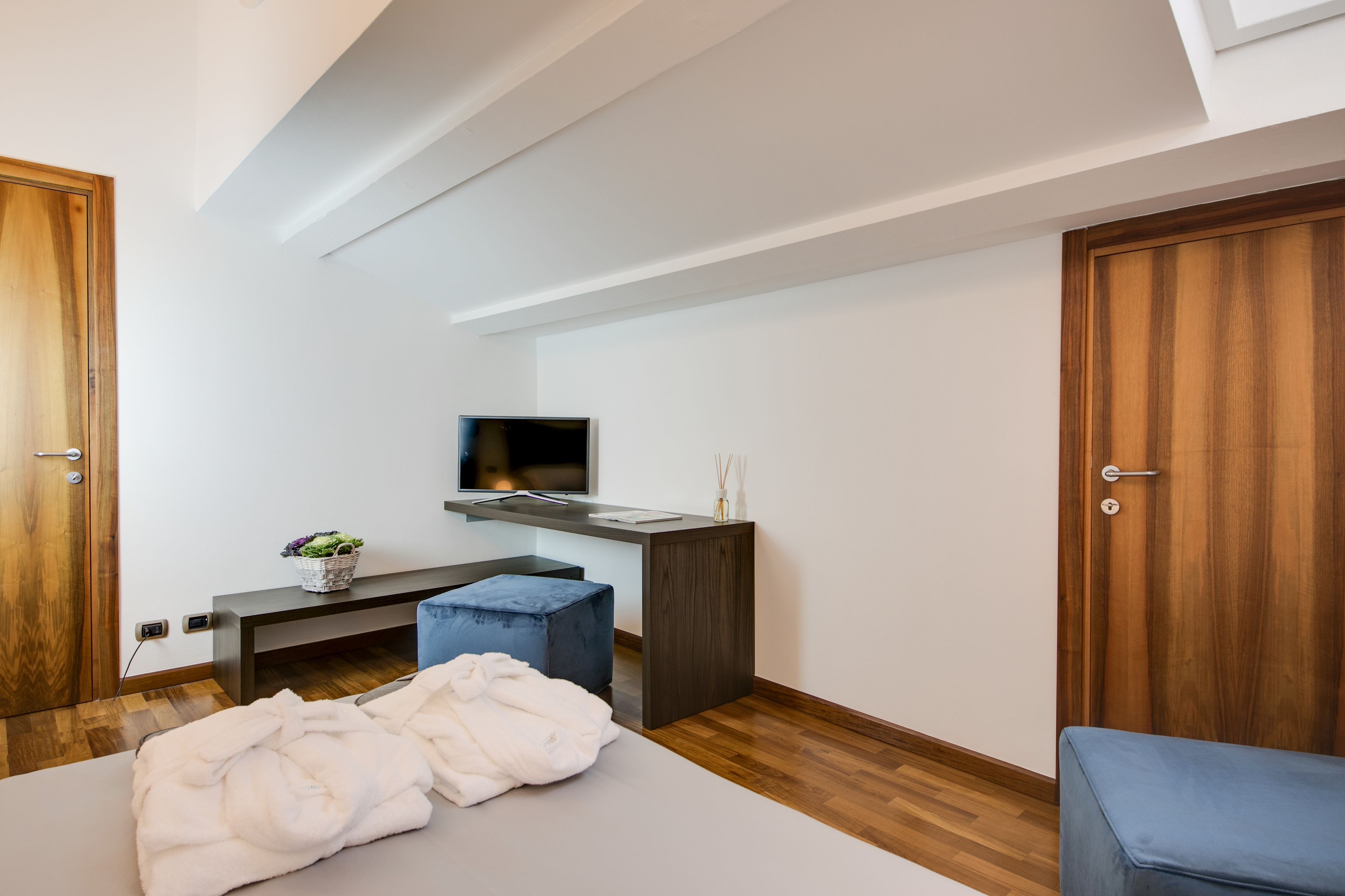 Une chambre moderne avec un plancher en bois, un lit recouvert d’une couverture blanche, un banc bleu, un bureau avec un téléviseur et une porte en bois.