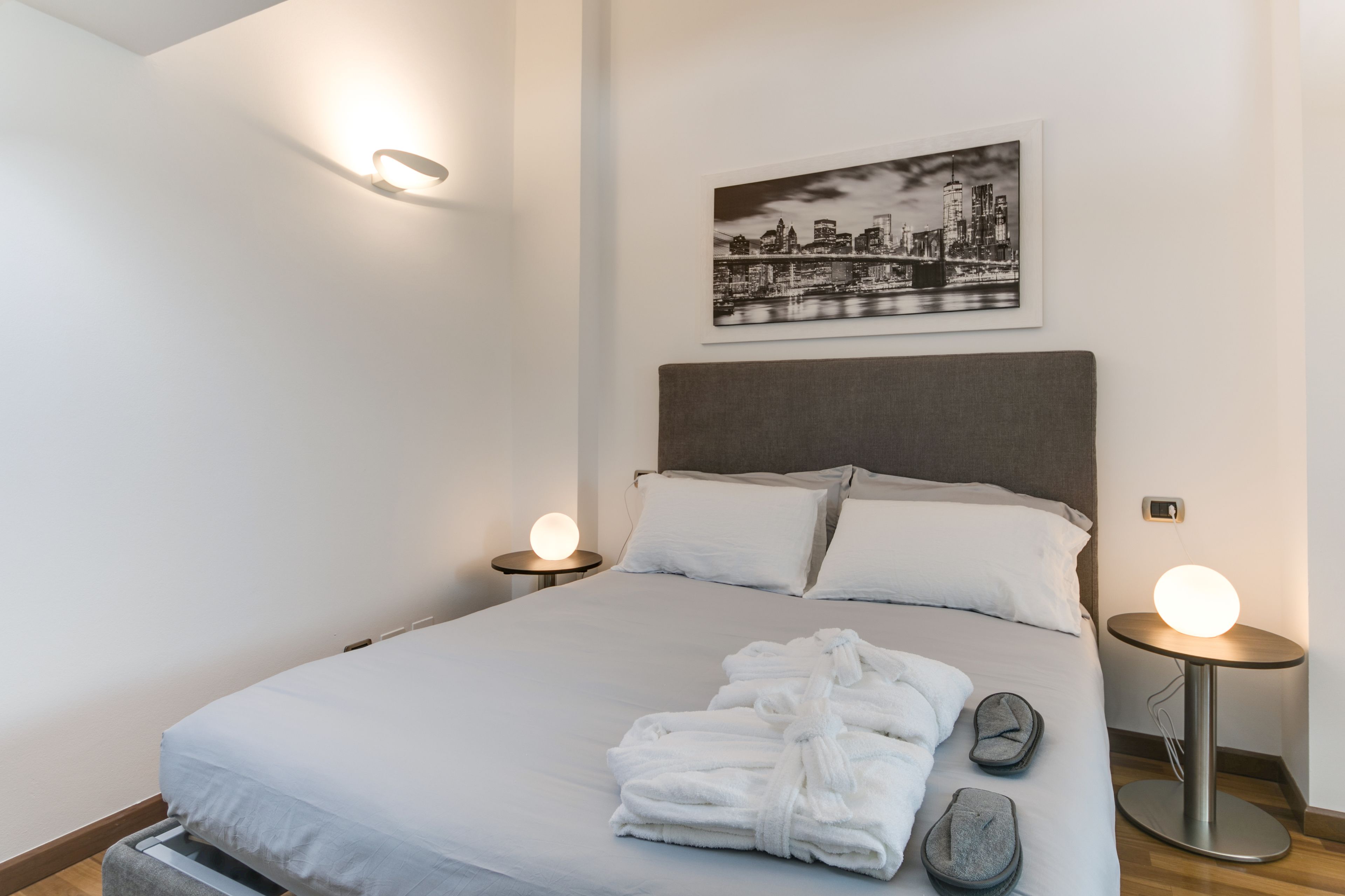 En snyggt bäddad säng med vita lakan och två kuddar, en inramad tavla med en stadsbild på väggen och två sängbord med runda lampor.