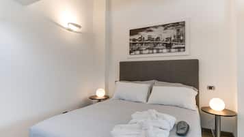En snyggt bäddad säng med vita lakan och två kuddar, en inramad tavla med en stadsbild på väggen och två sängbord med runda lampor.
