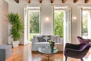 A modern living room with a white sofa, a glass coffee table, and purple chairs. There is a large potted plant and a vase with flowers on the table.