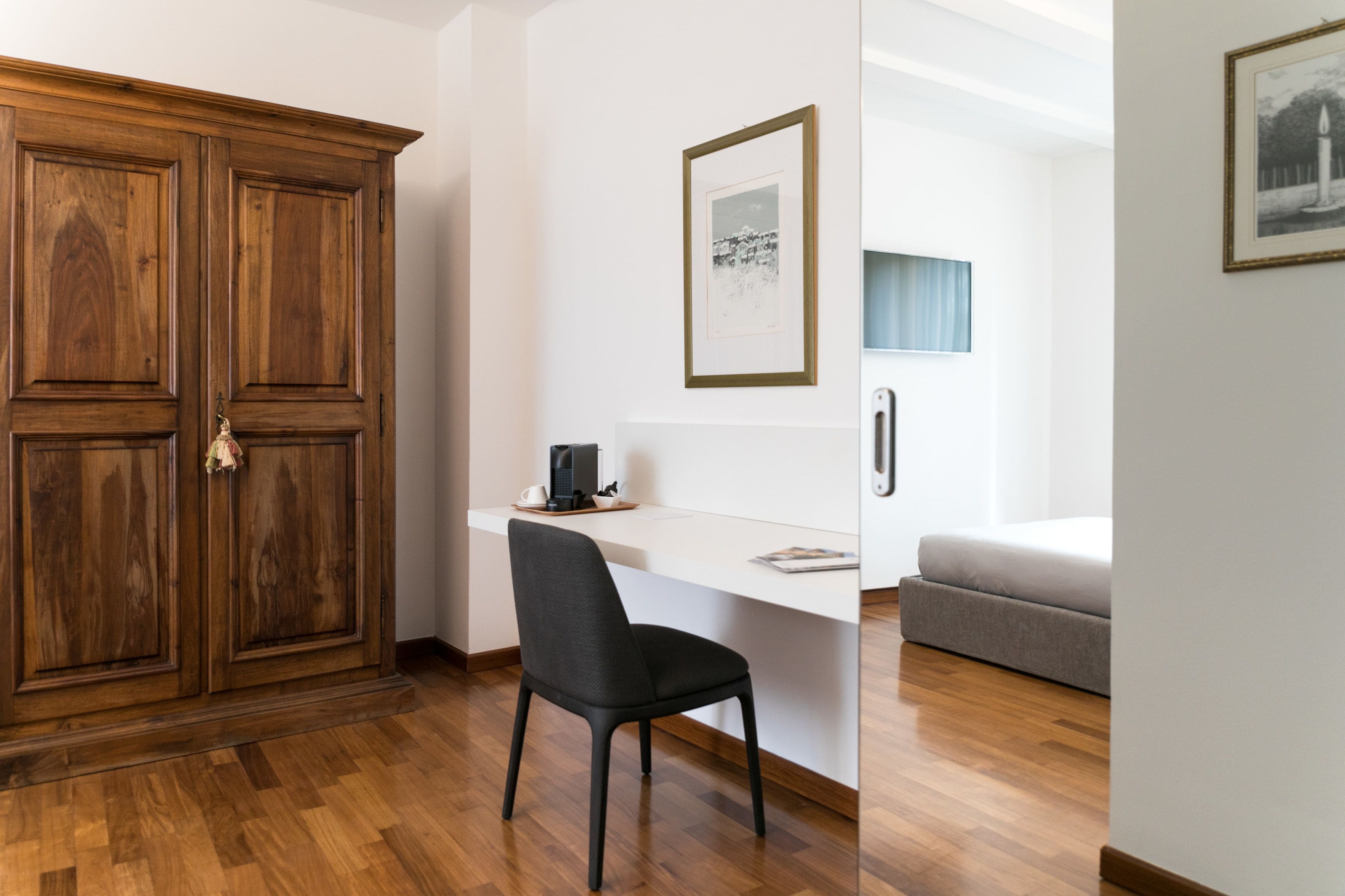 A room with a wooden wardrobe, a white desk with a chair, a framed picture, and a bed.