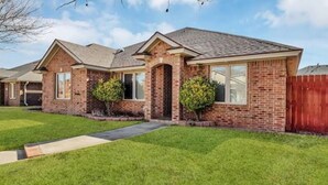 A single-story brick house with a front porch, a small front yard, and a red wooden fence. - Spark Getaways: The Shapiro (Lubbock)
