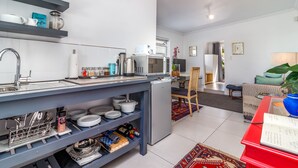 A modern kitchen with a built-in microwave, a coffee machine, and a small refrigerator. There is a dining area with a chair and a table, and a living room with a sofa and a lamp. - Bleu Cobalt Cottage (Franschhoek)