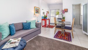 A modern living room with a grey sofa, blue cushions, a glass coffee table, and a newspaper on a blue ottoman. - Bleu Cobalt Cottage (Franschhoek)