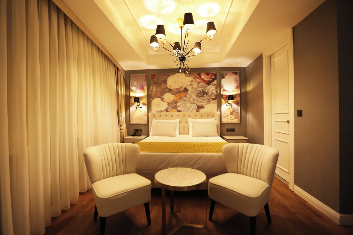 A hotel room with a bed, two armchairs, a small round table, a chandelier, and a floral wallpaper.