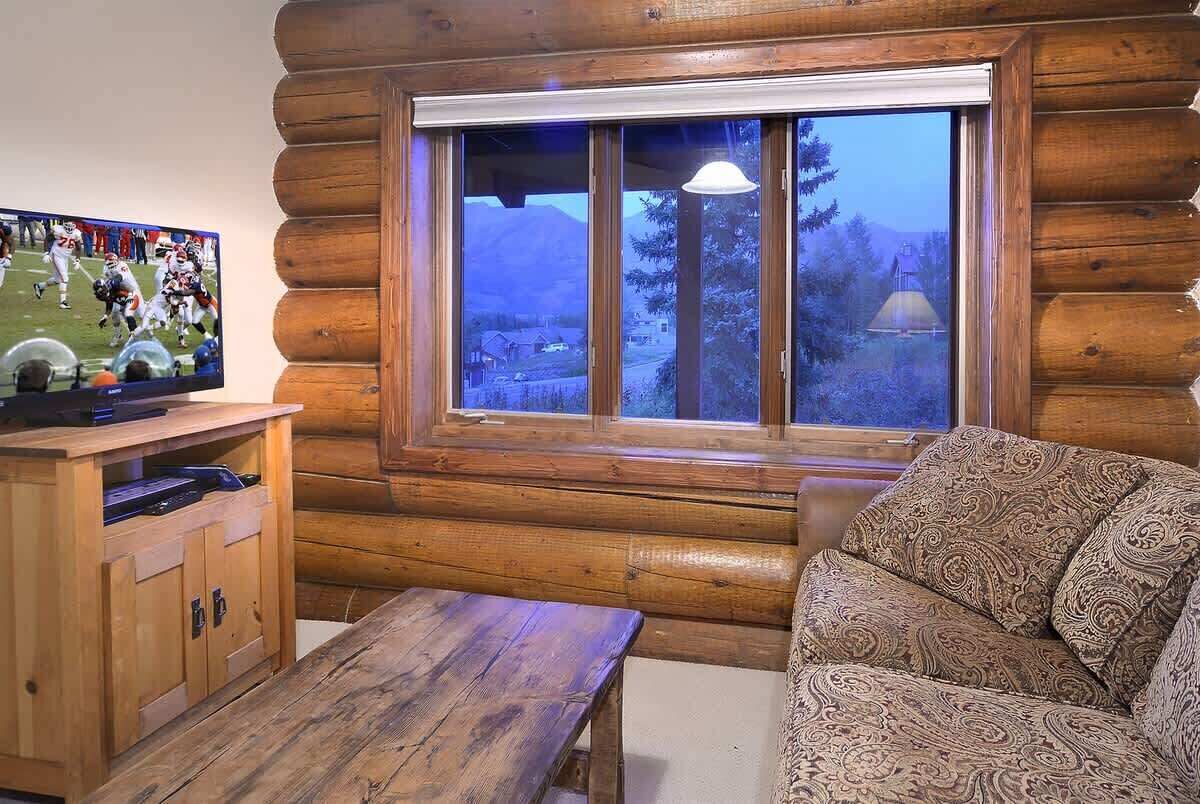 A log cabin interior with a wooden bench, a television on a wooden stand, and a window with a view of mountains and trees.