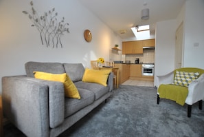 Cozy living space with a sofa, armchair, and dining area, featuring a modern kitchen with a wooden table and stainless steel oven.