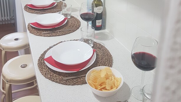 A dining table set with plates, glasses, and a bottle of wine, with a bowl of chips and a vase of flowers on the counter. - Appartamento Gradisca (Lecco)