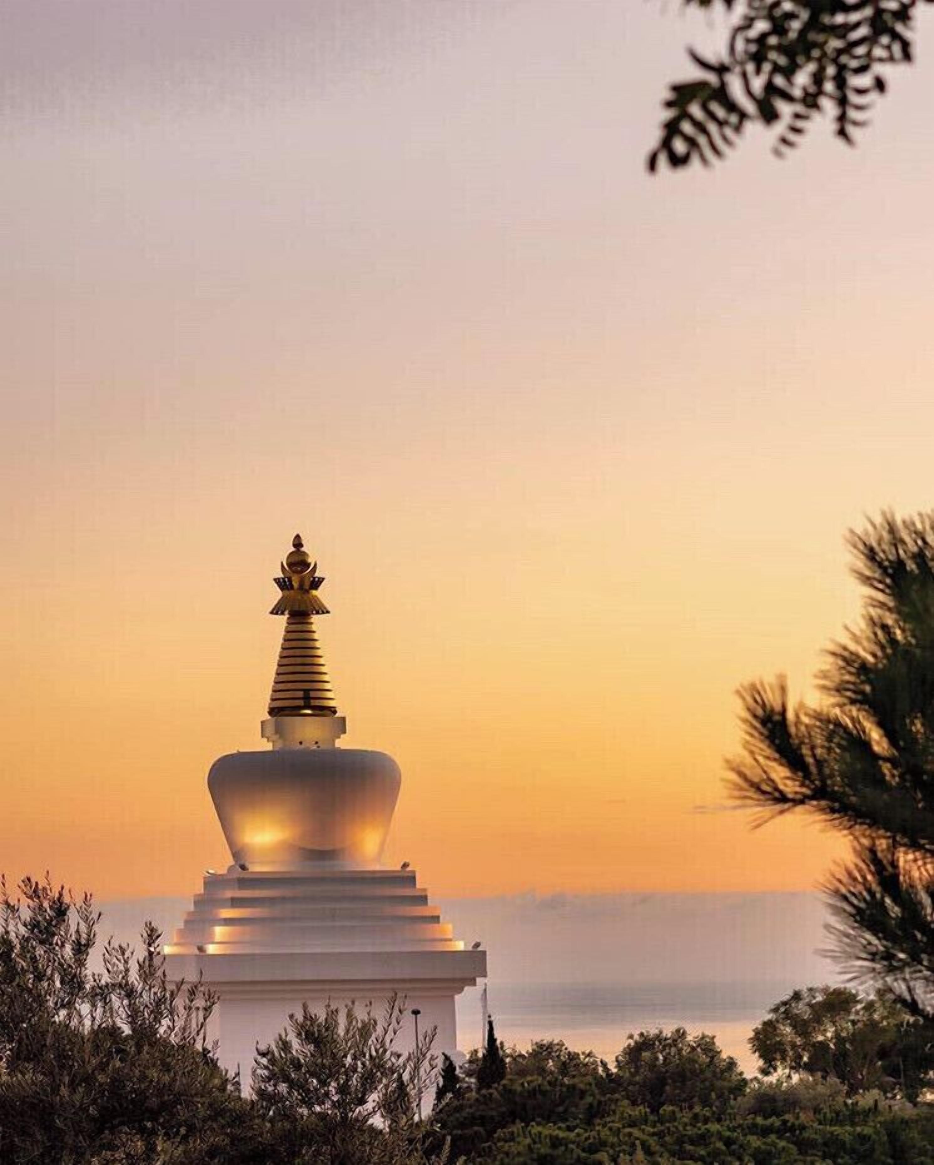 A white stupa with a golden spire, surrounded by greenery, against a sunset sky.