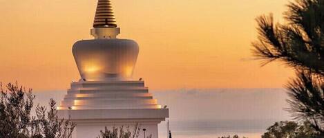 A white stupa with a golden spire, surrounded by greenery, against a sunset sky.