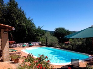 A rectangular swimming pool with a tiled deck, surrounded by a wooden fence and covered with a green canopy. There are lounge chairs and a hanging chair near the pool, and a stone building with a window to the left. - B&B Il Ciliegio (Manziana)