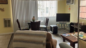 Een woonkamer met een bank, een eettafel met stoelen, een televisie en een raam met zonwering.
