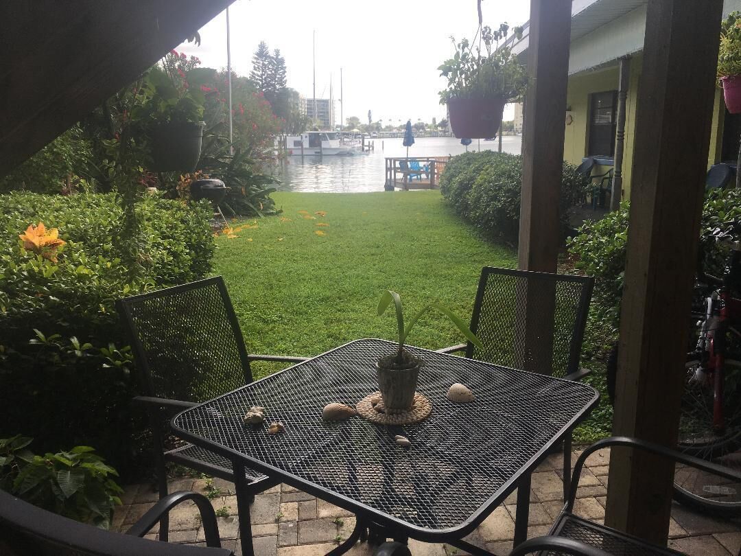 En uteplats med ett bord och stolar, en liten trädgård och utsikt över en kanal med båtar.