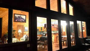 A cozy interior with wooden walls and large windows, a dining area with chairs, and a kitchen visible through the windows. - 2 Bedroom Hand Crafted Luxury Home with Jacuzzi & Fireplace (Volcano)