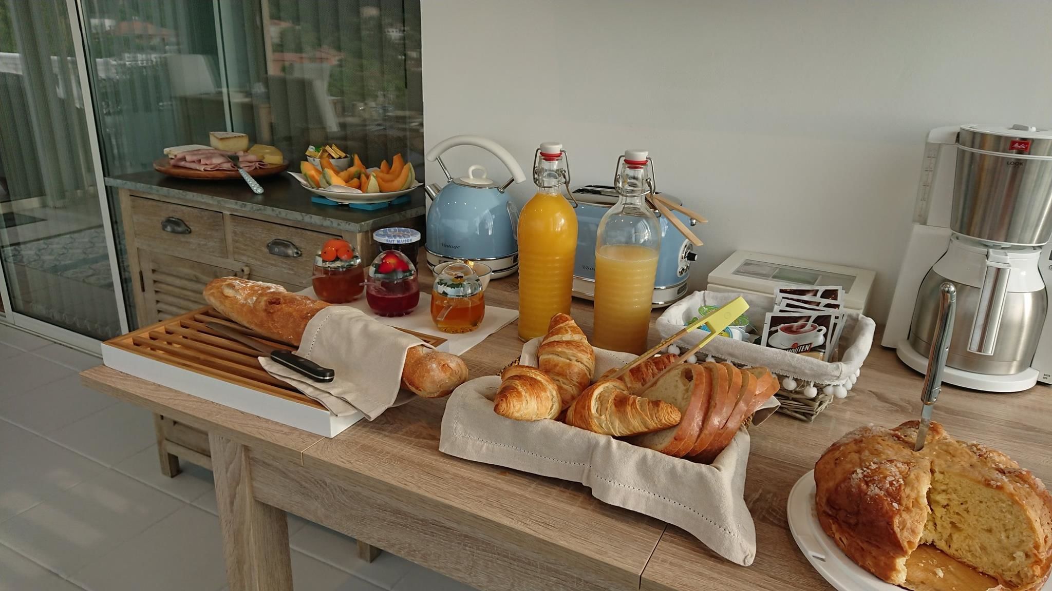 A breakfast table with croissants, bread, juice, and a coffee maker.