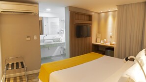 Habitación de hotel con cama, una silla, televisor, escritorio pequeño y baño visible a través de una puerta abierta.