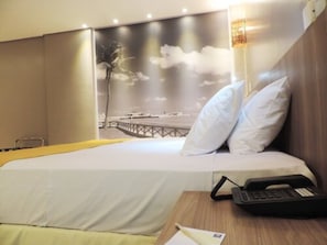 Quarto de hotel com uma cama, uma mesa de cabeceira com telefone e uma parede decorada com uma obra de arte cênica.