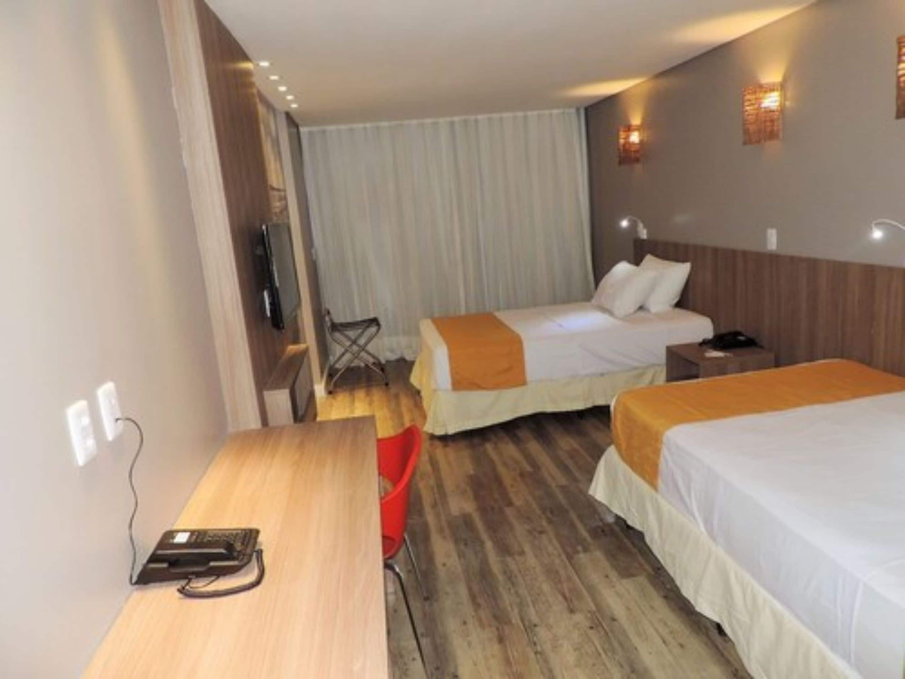 Quarto de hotel com duas camas, uma escrivaninha de madeira, uma cadeira e uma televisão.