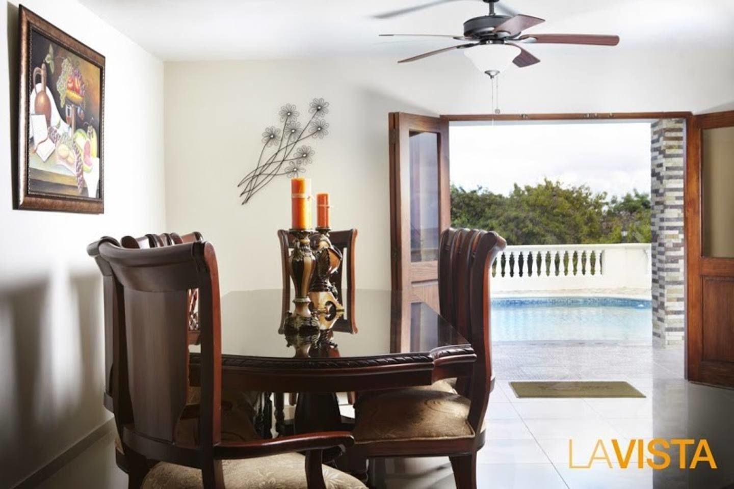 Un comedor con mesa y sillas, un centro decorativo y vistas a una piscina a través de una puerta abierta.
