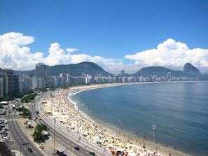 Un paisaje costero con una playa extensa, edificios urbanos y montañas al fondo.
