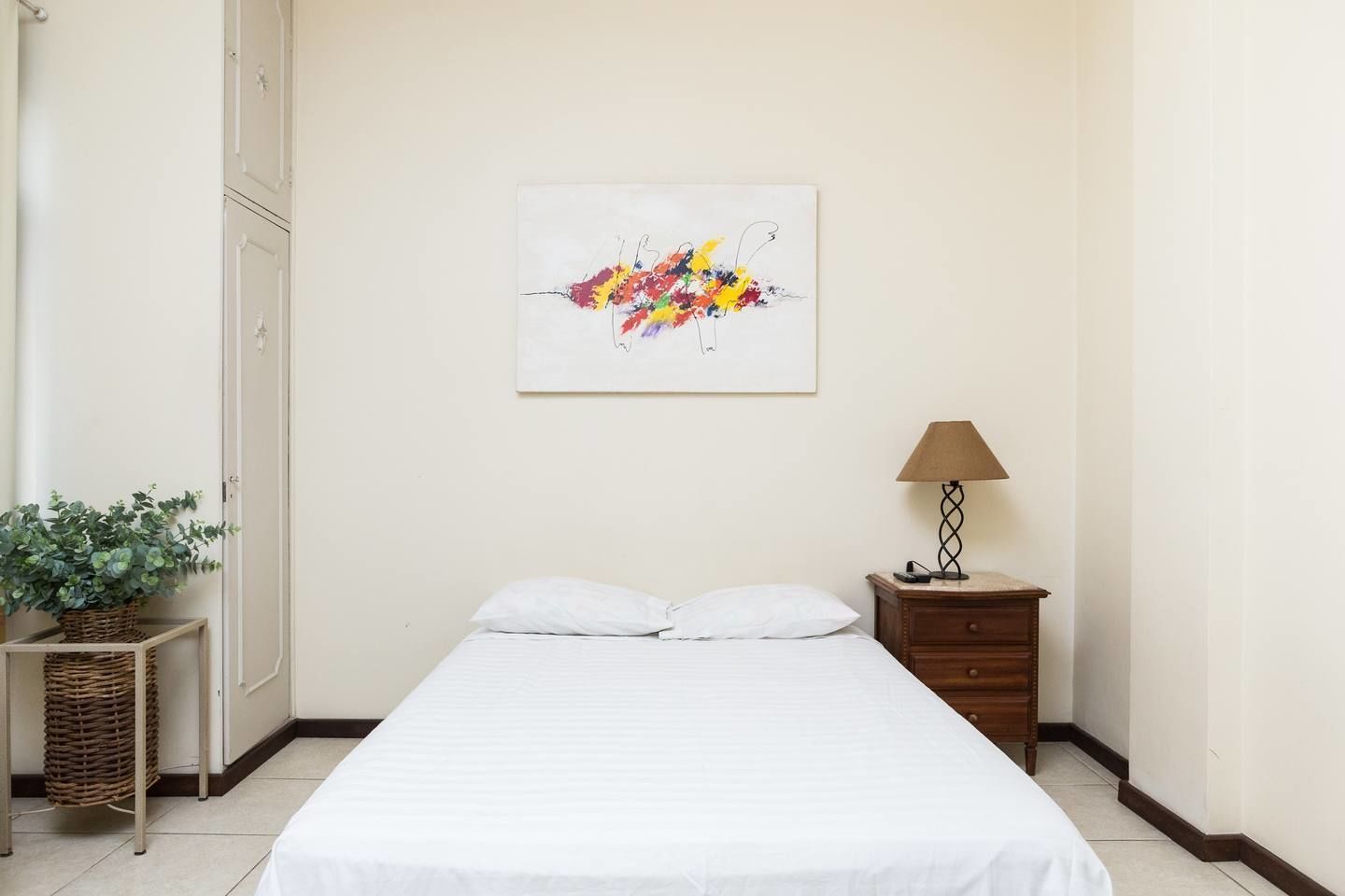 A bedroom with a bed, a nightstand, a lamp, a potted plant, and a wall-mounted abstract painting.
