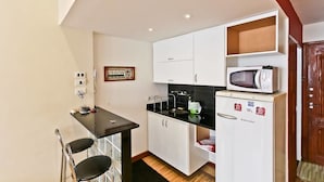 A compact kitchen with white cabinets, a black countertop, a microwave, and a refrigerator. There is a small table with two chairs and a framed picture on the wall. - Modern Studio by the Beach 103 Apartment 1 (Rio de Janeiro)