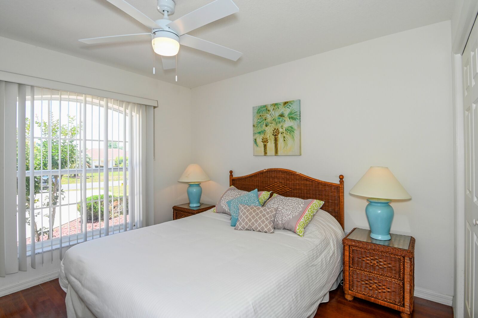 A bedroom with a large bed, two nightstands, a ceiling fan, a window with blinds, and a painting on the wall.
