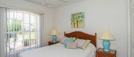 A bedroom with a large bed, two nightstands, a ceiling fan, a window with blinds, and a painting on the wall.