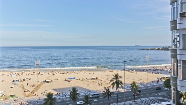 Une vue sur la plage depuis un immeuble de grande hauteur, avec un ciel dégagé, des palmiers et une plage animée, parsemée de parasols et de personnes.