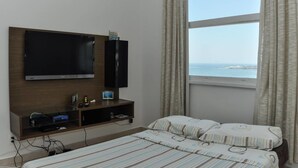 Une chambre d’hôtel avec un lit, un téléviseur fixé au mur, une petite étagère avec quelques objets et une fenêtre donnant sur la mer.