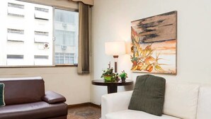 A living room with a brown leather sofa, a white sofa, a painting on the wall, a lamp on a side table, and a window with blinds. - Wizzie's 3 Bedoom Beach Pad - 304 Apartment 3 (Rio de Janeiro)