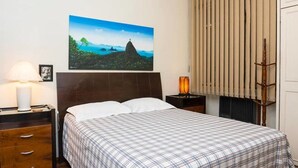 A hotel room with a bed, a nightstand with a lamp, a picture of a landscape, and a window with blinds. - Wizzie's 3 Bedoom Beach Pad - 304 Apartment 3 (Rio de Janeiro)