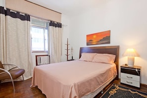 A bedroom with a bed, a bedside table with a lamp, a chair, a window with curtains, a painting on the wall, and a radiator. - Wizzie's 3 Bedoom Beach Pad - 304 Apartment 3 (Rio de Janeiro)