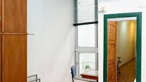 Een compacte kamer met een houten kledingkast, een bureau met stoel, een raam en een spiegel die een gang weerspiegelt.