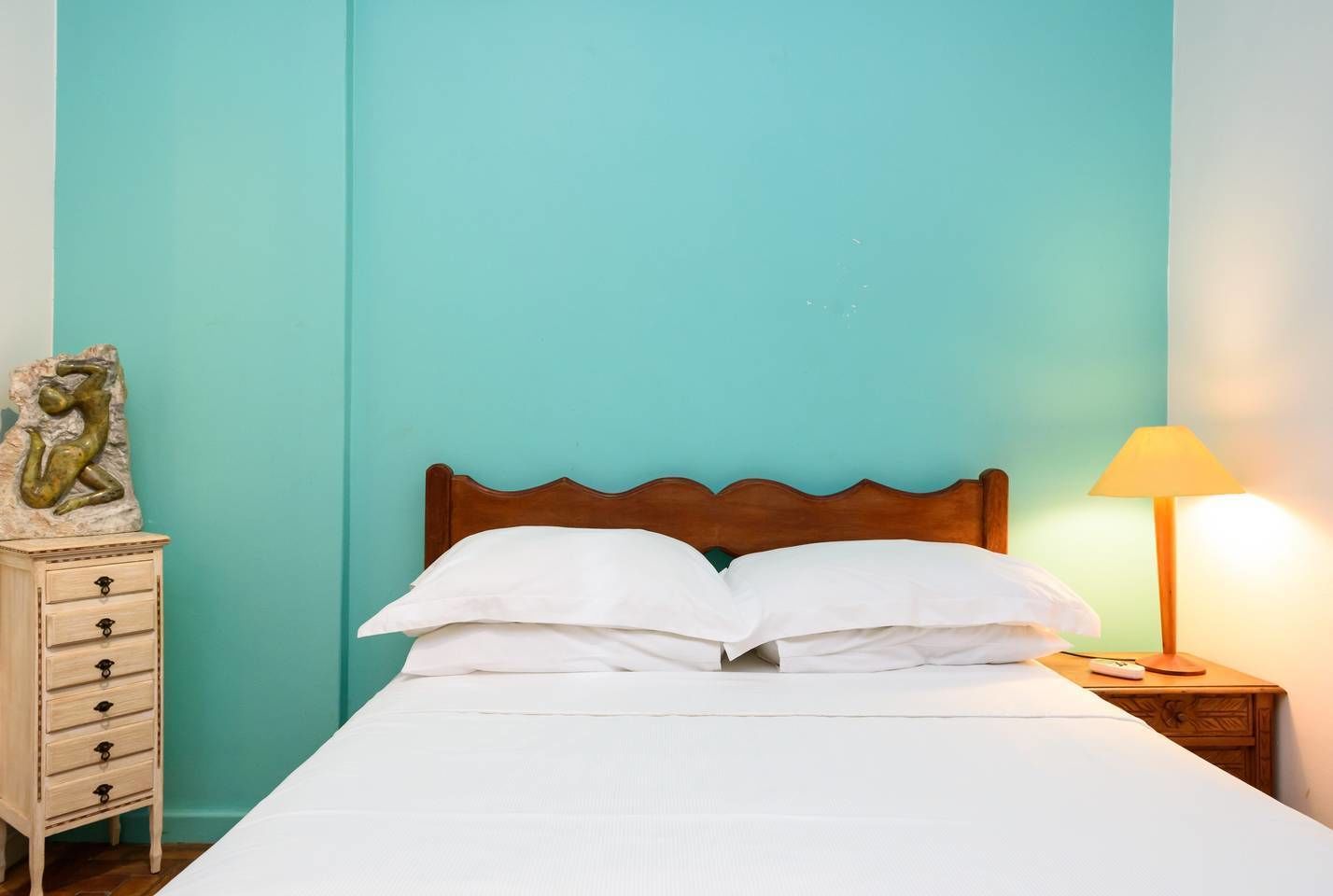 Un lit bien fait, avec du linge de lit blanc, dans une pièce aux murs de couleur turquoise et une table de chevet en bois sur laquelle se trouve une lampe.