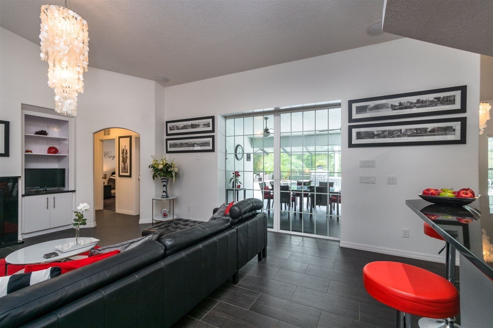 A modern living room with a black leather sofa, a glass coffee table, and a red bar stool. There is a dining area with a glass door leading to a patio.