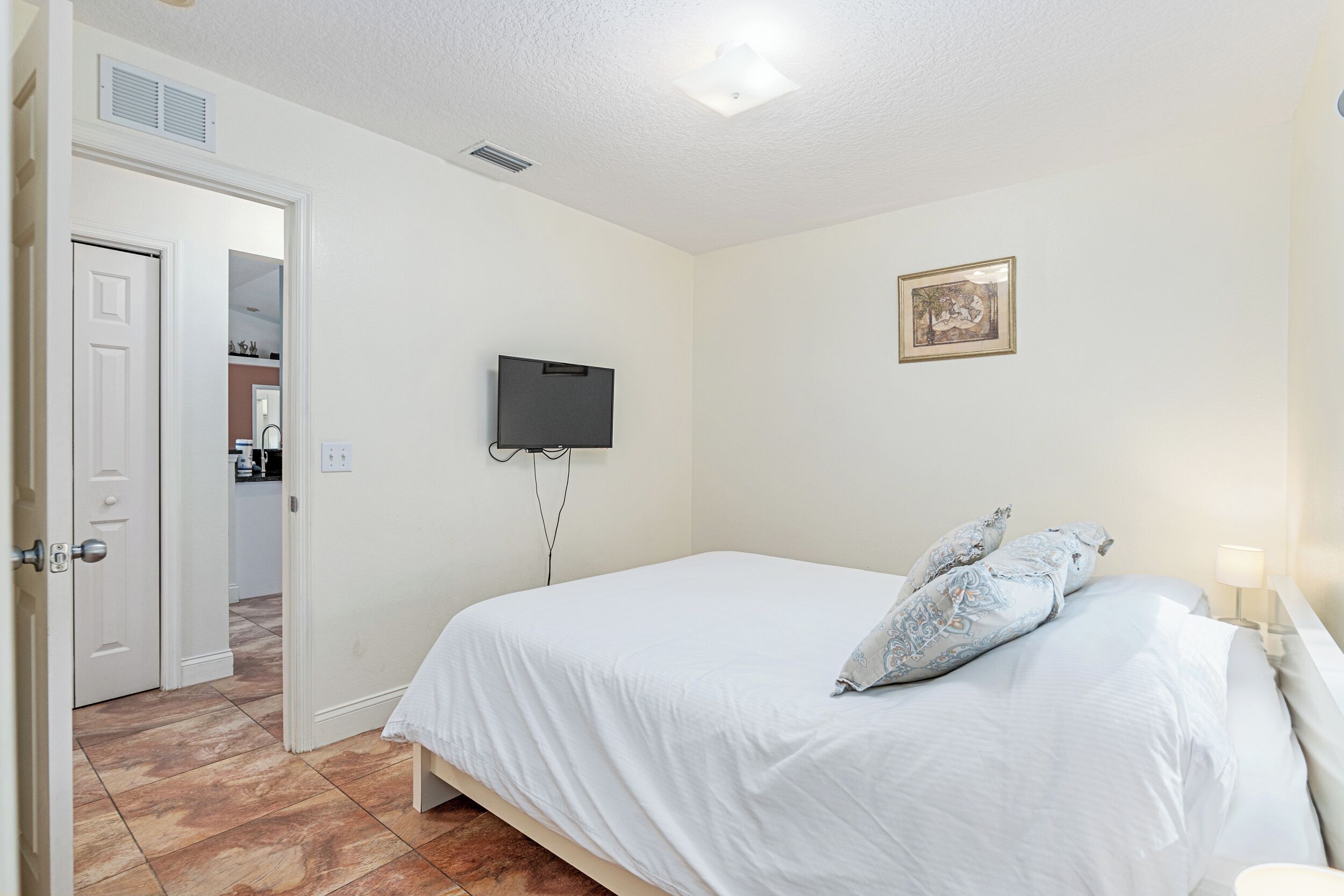 Een slaapkamer met een bed, een aan de muur bevestigde televisie, een deur naar een andere kamer en een klein lijstje met een afbeelding aan de wand.