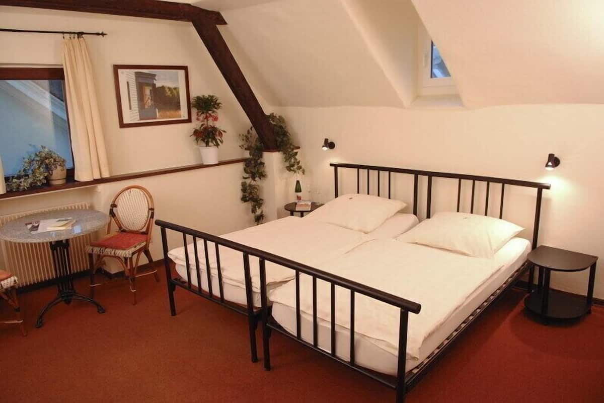 Um quarto com cama, uma cadeira, uma mesinha e uma janela com cortinas.