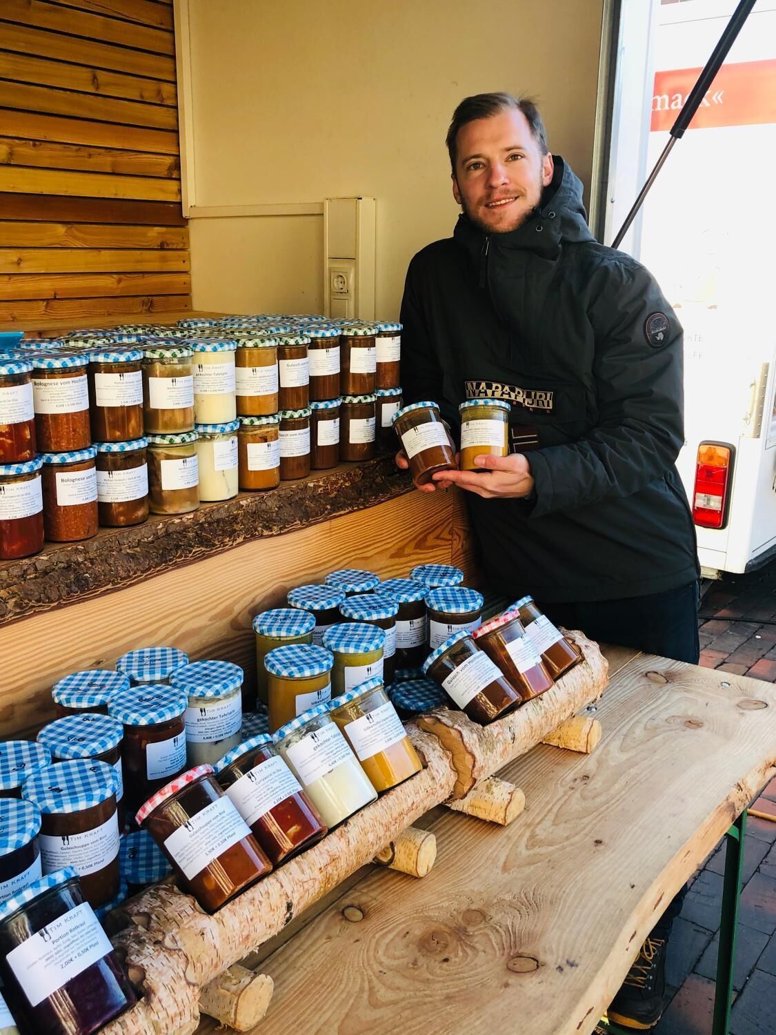 Eine Person hält ein Einmachglas auf einem Marktstand, wo verschiedene Gläser mit Konserven auf Holzböden und einem Tisch ausgestellt sind.