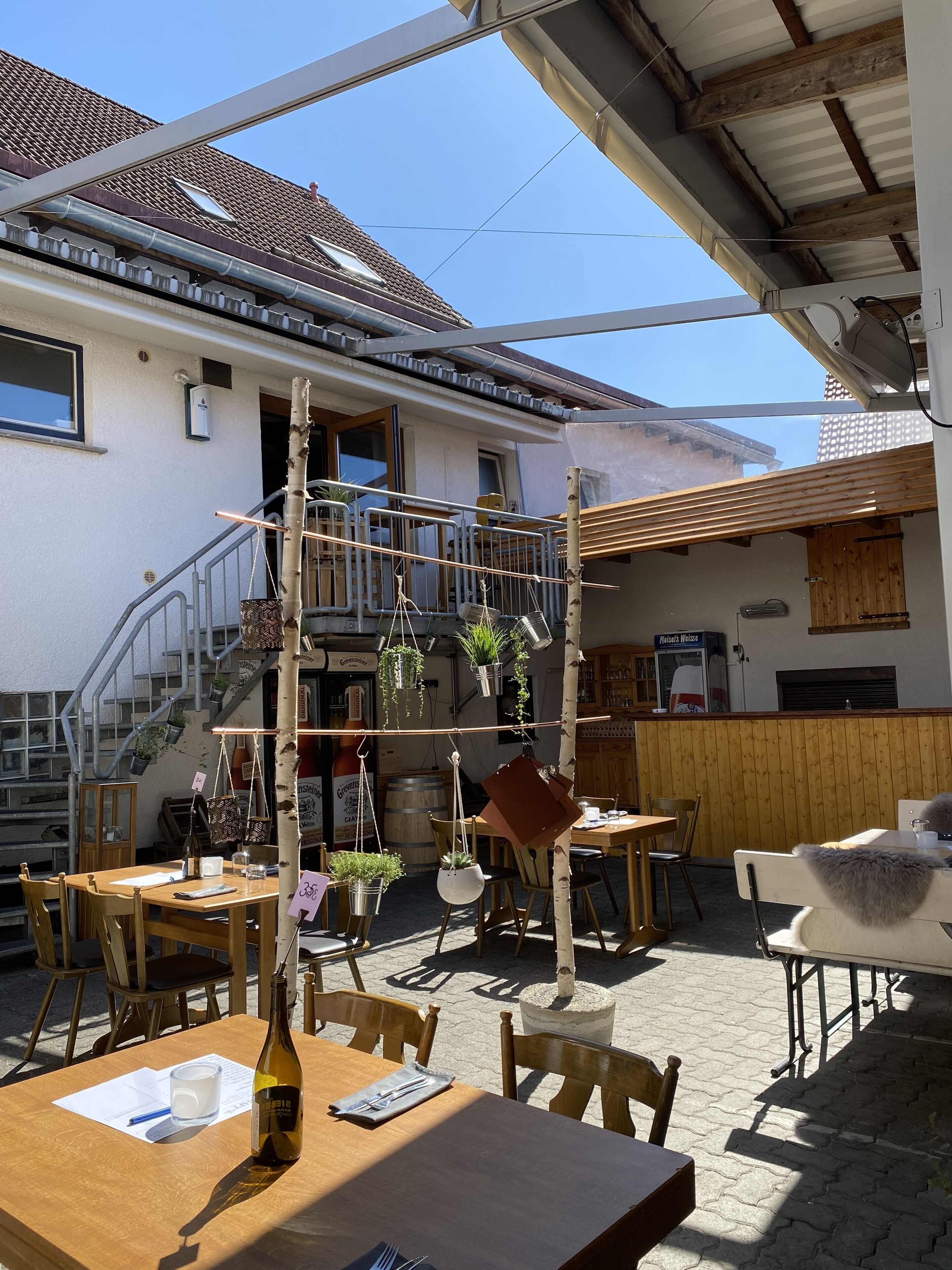 Un café en plein air avec des tables et des chaises en bois, un escalier menant à un balcon et des plantes suspendues.