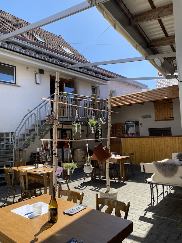 An outdoor café with wooden tables and chairs, a staircase leading to a balcony, and hanging plants.