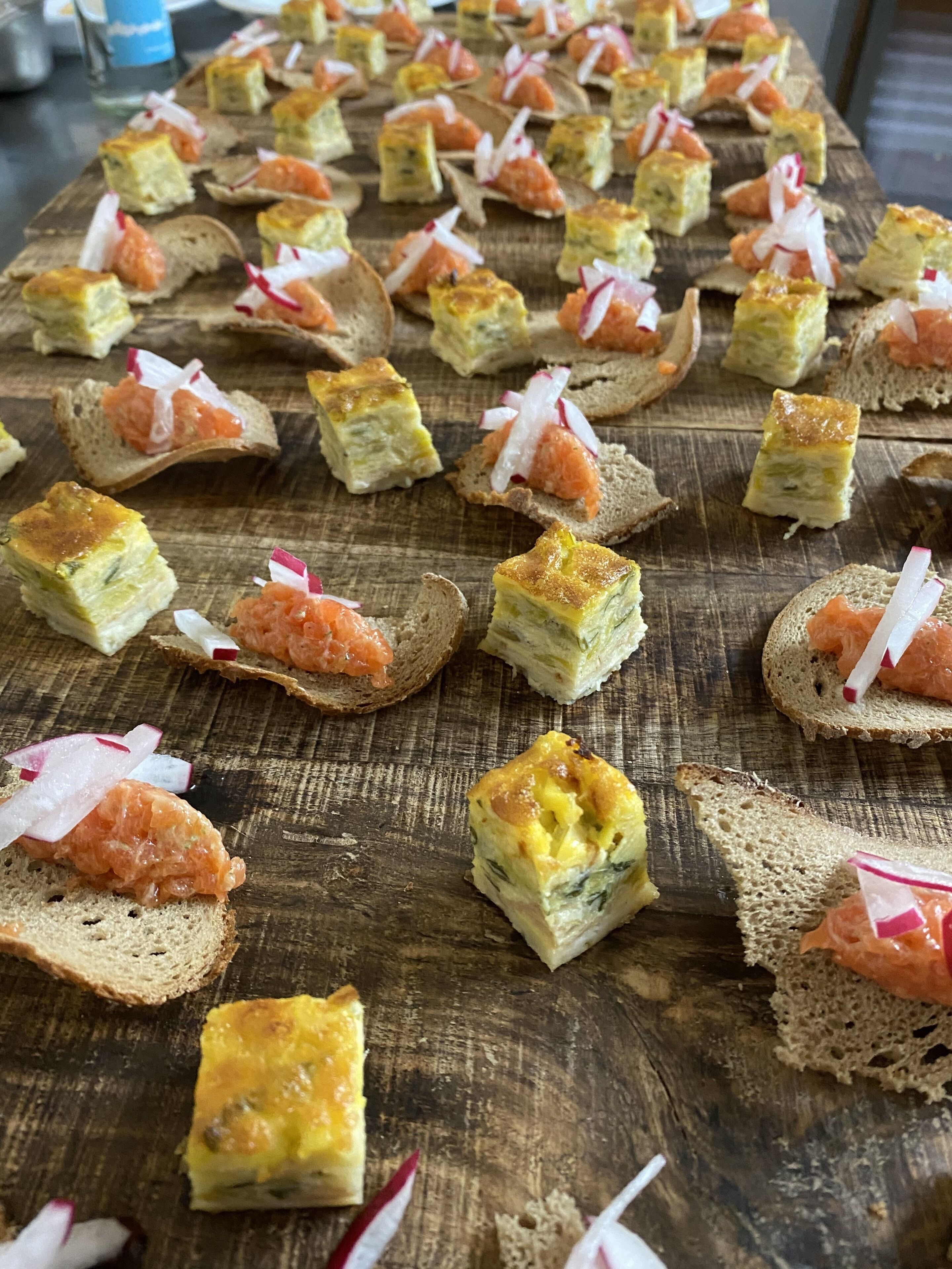 En træplanke med forskellige appetizers, heriblandt røget laks og en tærteagtig ret, anrettet på brødskiver.