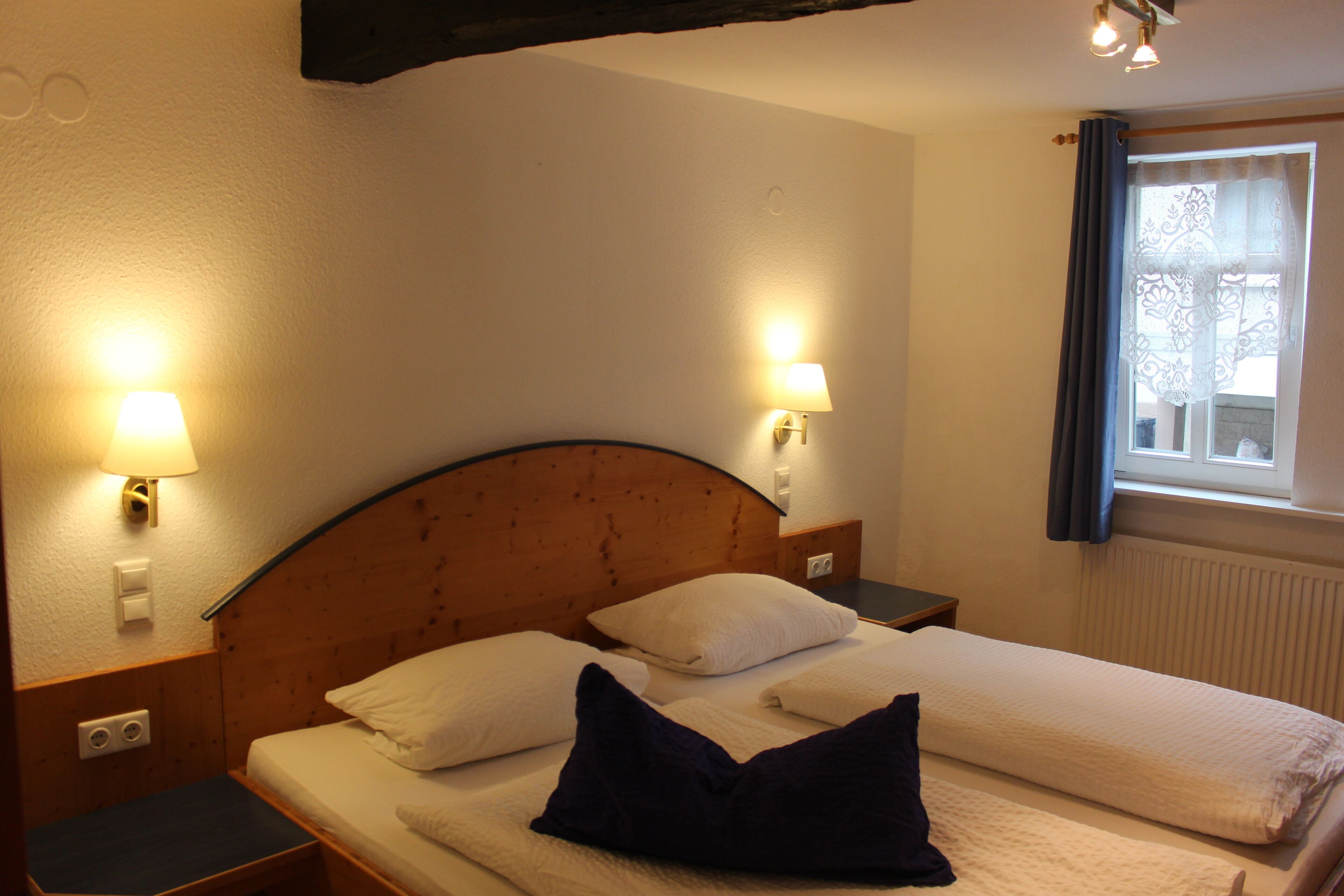 A hotel room with a wooden headboard, two bedside tables with lamps, a window with curtains, and a bed with pillows and a blanket.