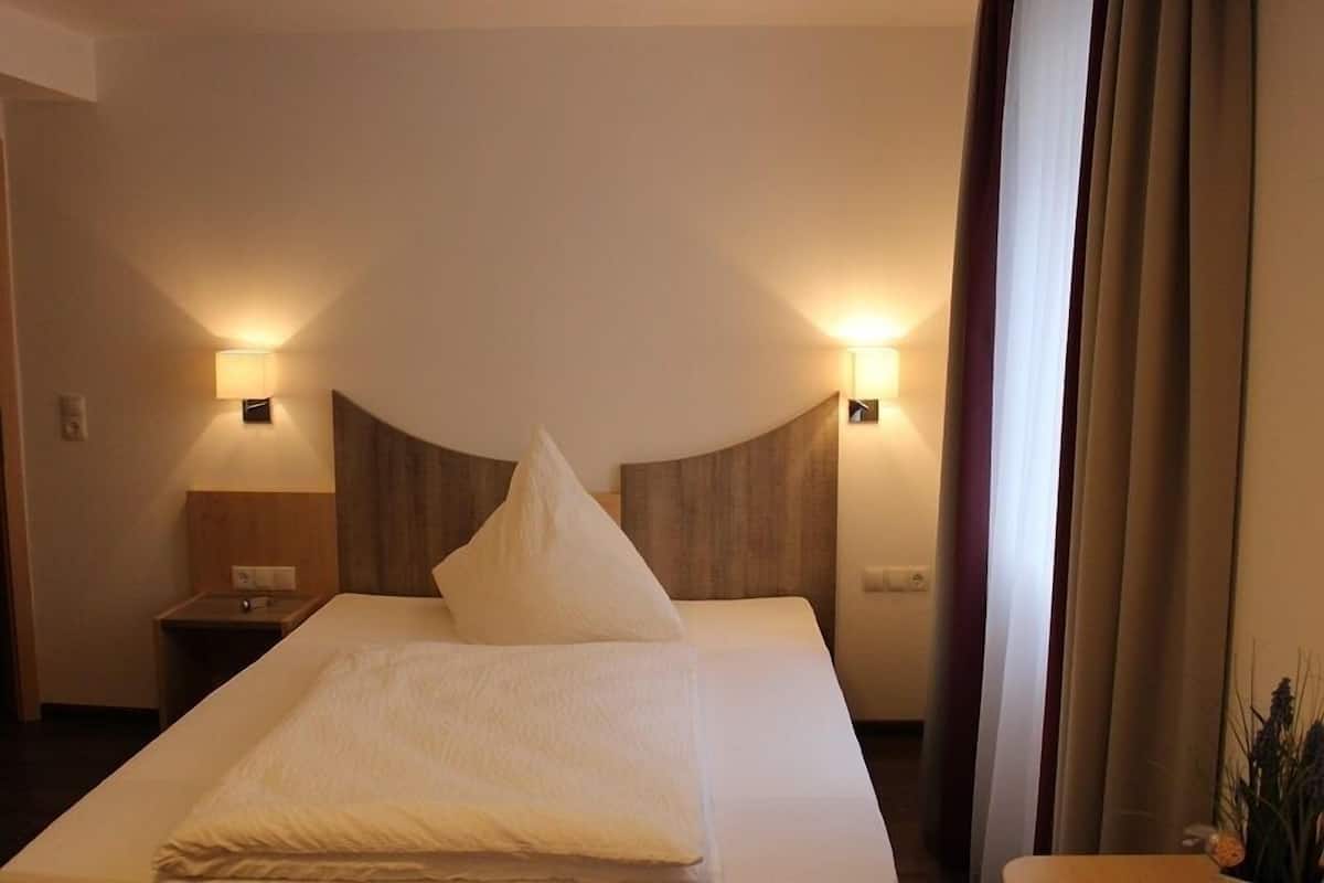 A hotel room with a bed, two bedside lamps, a nightstand, and a window with curtains.