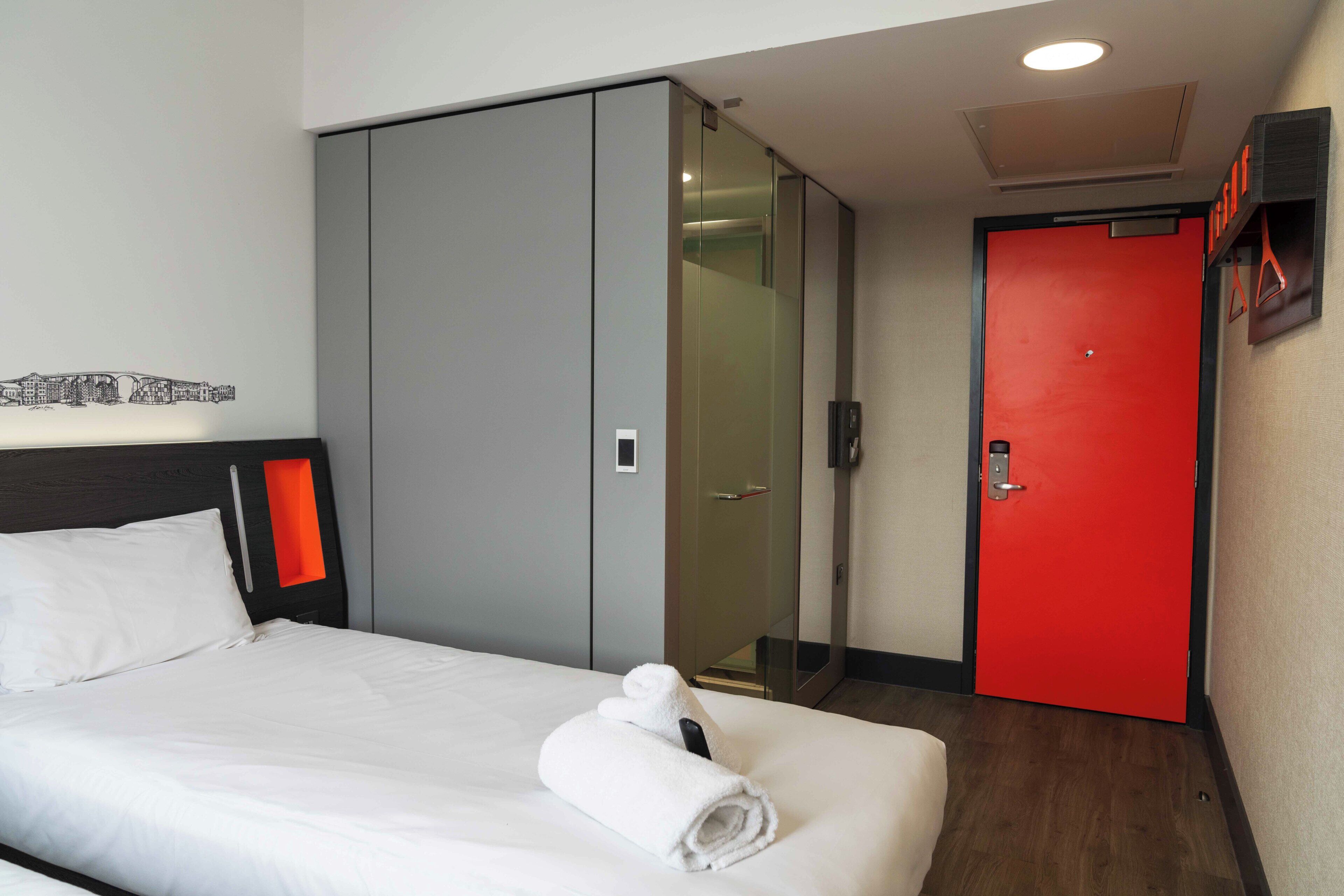 A hotel room with a bed, a wardrobe, a glass door, and a red door.