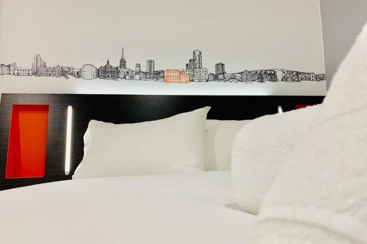 Ein Hotelzimmer mit Bett, weißer Bettwäsche, einem Wandgemälde mit Stadtansicht und einer roten Akzentwand.