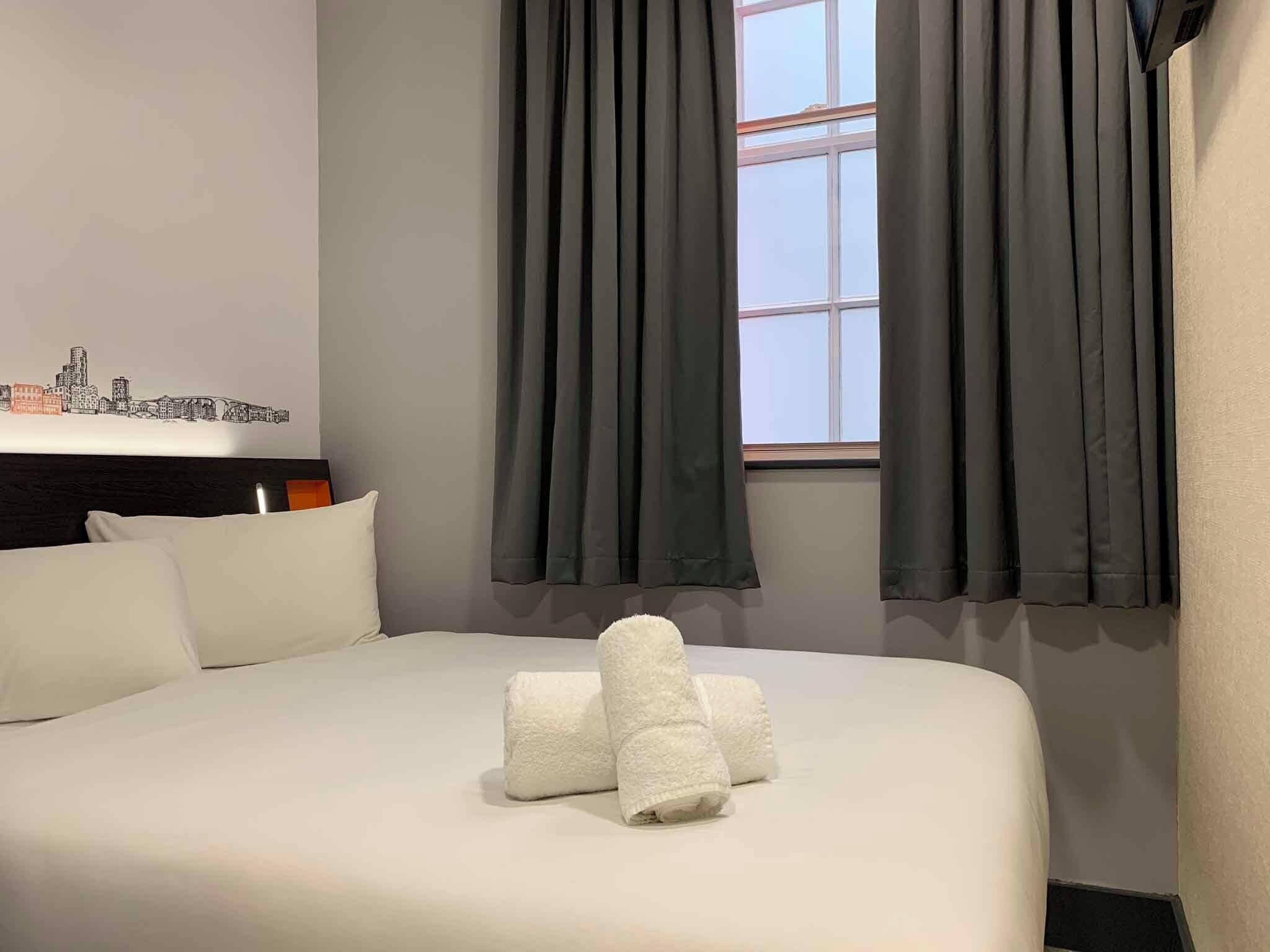 A hotel room with a bed, pillows, a window with curtains, and a wall with a cityscape artwork.