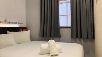 Quarto de hotel com uma cama, almofadas, janela com cortinas e um quadro com uma vista urbana.