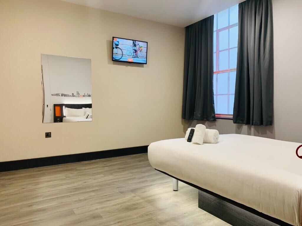 A hotel room with a bed, a TV mounted on the wall, a window with curtains, and a mirror.