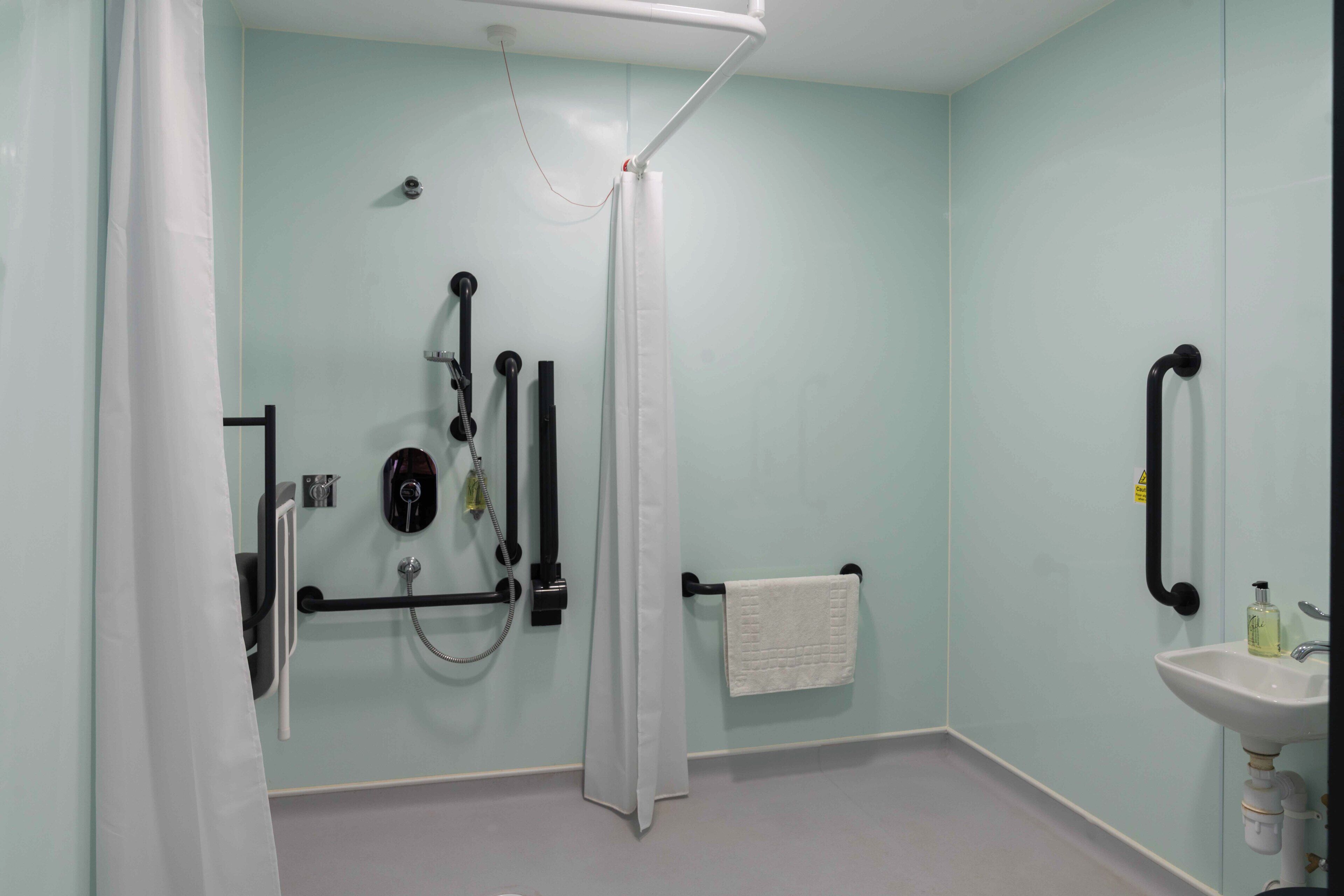 Une salle de bain équipée d’une douche, de barres d’appui, d’un porte-serviettes et d’un lavabo.